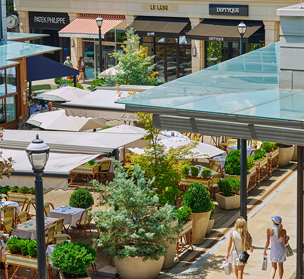 Shoppers walk through an open-air retail village with outdoor dining and luxury storefronts.