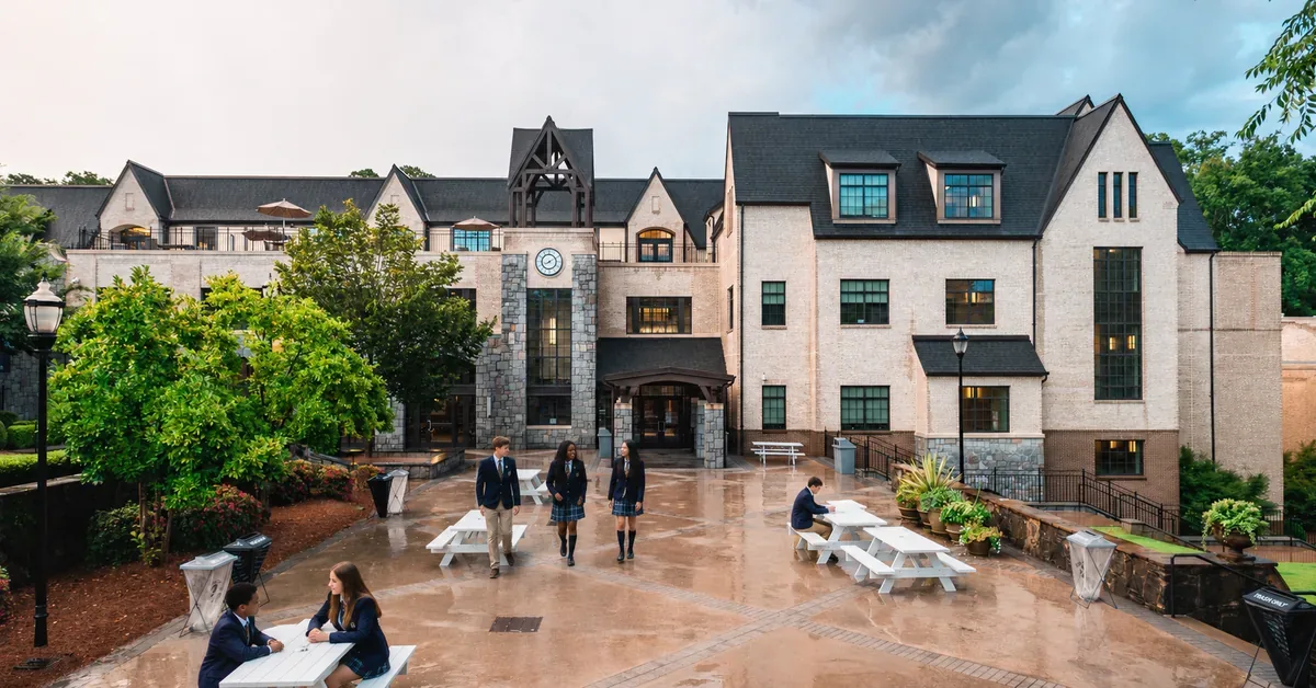 Students in uniforms gather in the wet courtyard of an elegant Tudor-style private school campus featuring stone facades and dark timber accents.
